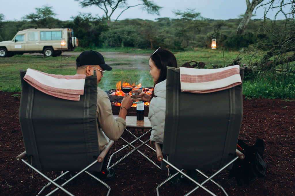 Couple having sundowner drinks in camp chairs with the safari vehicle behind